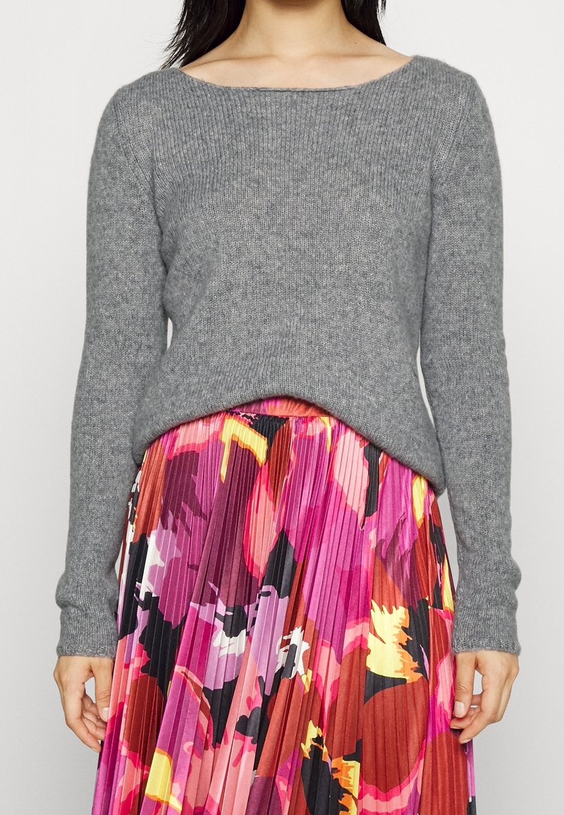 Grijze gebreide trui met een brede, boothals en lange mouwen, gecombineerd met een kleurrijke geplooide bloemenrok in roze- en oranje tinten.