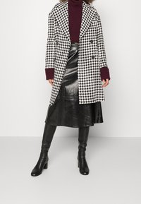 Svartvitt houndstooth-mönstrat överplagg med överdimensionerad krage, parat med en vinröd polotröja, svart läderkjol och knähöga svarta stövlar.