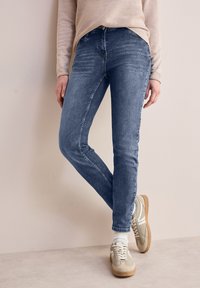 Jean en denim bleu slim avec légère décoloration, cinq poches et passants de ceinture, associés à des baskets de couleur claire et des chaussettes blanches.
