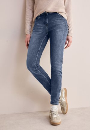 Schmal geschnittene blaue Jeans aus Denim mit leichtem Verblassen, fünf Taschen und Gürtelschlaufen, kombiniert mit hellen Sneakers und weißen Socken.