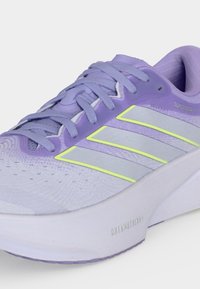 Chaussure de sport lilas et blanche avec laçage à l’avant, trois bandes bordées de jaune sur le côté et une semelle blanche épaisse marquée « Dreamstrides ».
