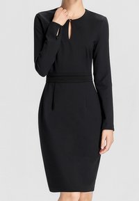 Femme portant une robe noire ajustée à manches longues, longueur genou, avec un col en trou de serrure et une taille cintrée.