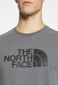 Camiseta de algodón gris con un gran logo negro de "The North Face" en el frente. Cuello redondo, mangas cortas, textura suave.