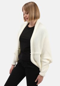 Frau mit kurzem blonden Haar, die eine weiße gestrickte offene Strickjacke über einem schwarzen Shirt und schwarzen Hosen vor einem weißen Hintergrund trägt.