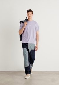 Helllila T-Shirt, lockere Passform; kombiniert mit Jeans im Patchworkdesign in Dunkel- und Hellblau; Modell hält eine Jacke; neutraler Hintergrund.