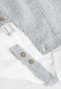 Chemise à col blanc avec une superposition en tissu tricoté gris et blanc texturé, ornée de boutons ronds marron avec du texte.
