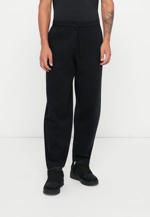 Black cotton jogger pants with an elastic waistband and drawstring, featuring side pockets and a relaxed fit. Worn with black sneakers.