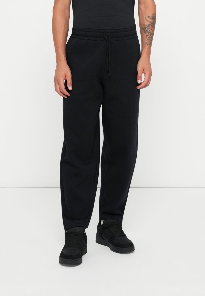 Black cotton jogger pants with an elastic waistband and drawstring, featuring side pockets and a relaxed fit. Worn with black sneakers.