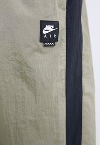 Nike Air hlače, ki se ponašajo s lahko, teksturirano tkanino v olivno zeleni in temno modri barvi. Vključujejo izrazit črn logotip.
