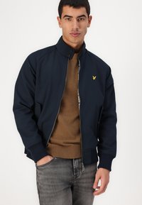 Giacca da bombardiere blu navy con colletto alto, polsini a costine e dettaglio del logo dorato. Indossata sopra un maglione marrone, abbinata a jeans grigi.