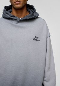 Grijze oversized hoodie met een textuurafwerking. Heeft een donkergrijze binnenkap en zwarte geborduurde tekst "Stay Moment" op de borst.
