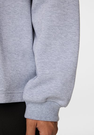 Zbliżenie na ramię osoby noszącej jasnoszary, długi rękaw bluzy z prążkowanymi mankietami, ręka swobodnie opuszczona wzdłuż ciała na białym tle.