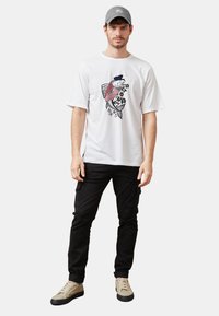 Camiseta blanca con estampado gráfico de un pez, pantalones cargo negros y zapatillas beige. También incluye una gorra gris con un logo.