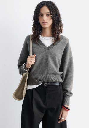 Jeune femme aux cheveux bouclés porte un pull gris à col en V, un pantalon noir, porte un sac à épaule beige et porte des bracelets et des bagues sur fond blanc.