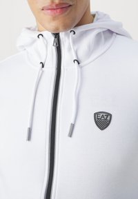 Felpa bianca con zip, zip nera, cordini e badge del logo EA7 sul petto. Tessuto liscio con un design aderente.