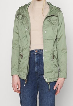 Femme portant une veste à capuche vert clair sur un pull à col roulé beige et un jean bleu, debout devant un fond uni.