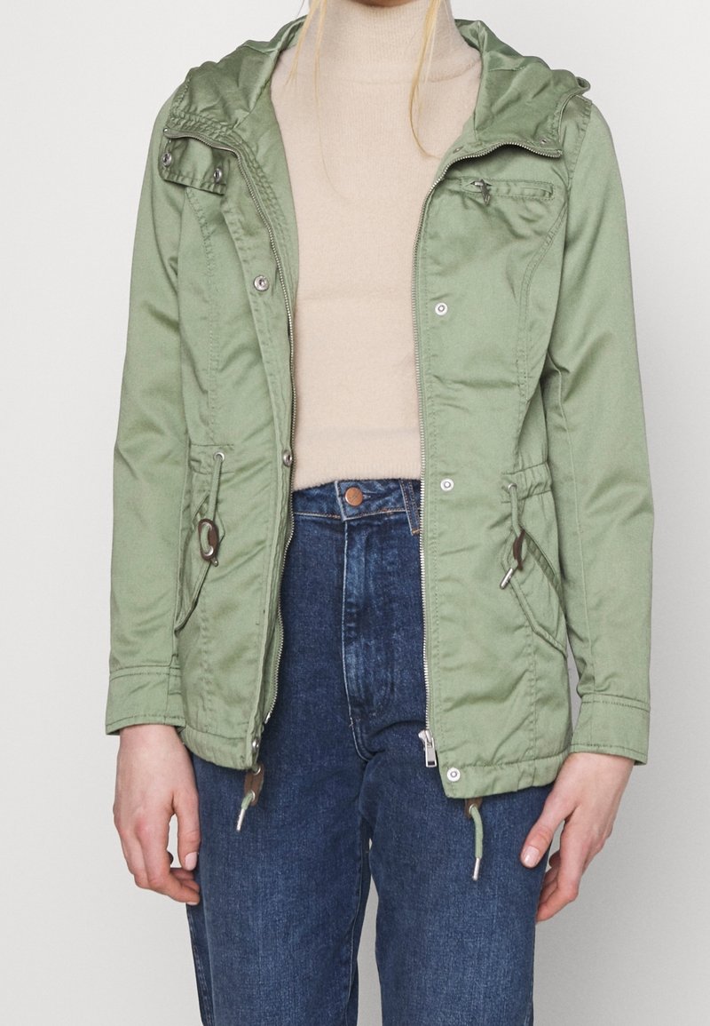 Femme portant une veste à capuche vert clair sur un pull à col roulé beige et un jean bleu, debout devant un fond uni.