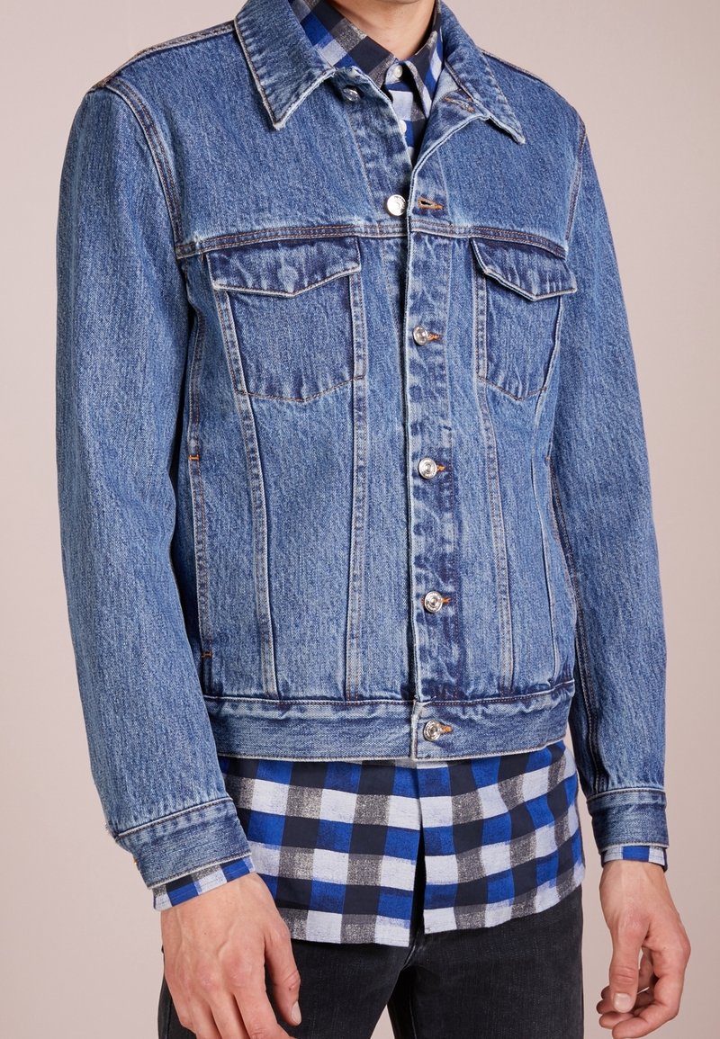 Mężczyzna w niebieskiej dżinsowej kurtce na tle niebiesko-czarno-białej kraciastej koszuli i czarnych spodni, na gładkim tle.