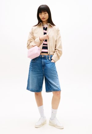 Jauna sieviete valkā bēša krāsas žaketi, svītrainu kreklu, džinsa šortus līdz ceļiem, baltus kedus, baltas zeķes un rozā plecu somiņu, stāv ar roku kabatā.