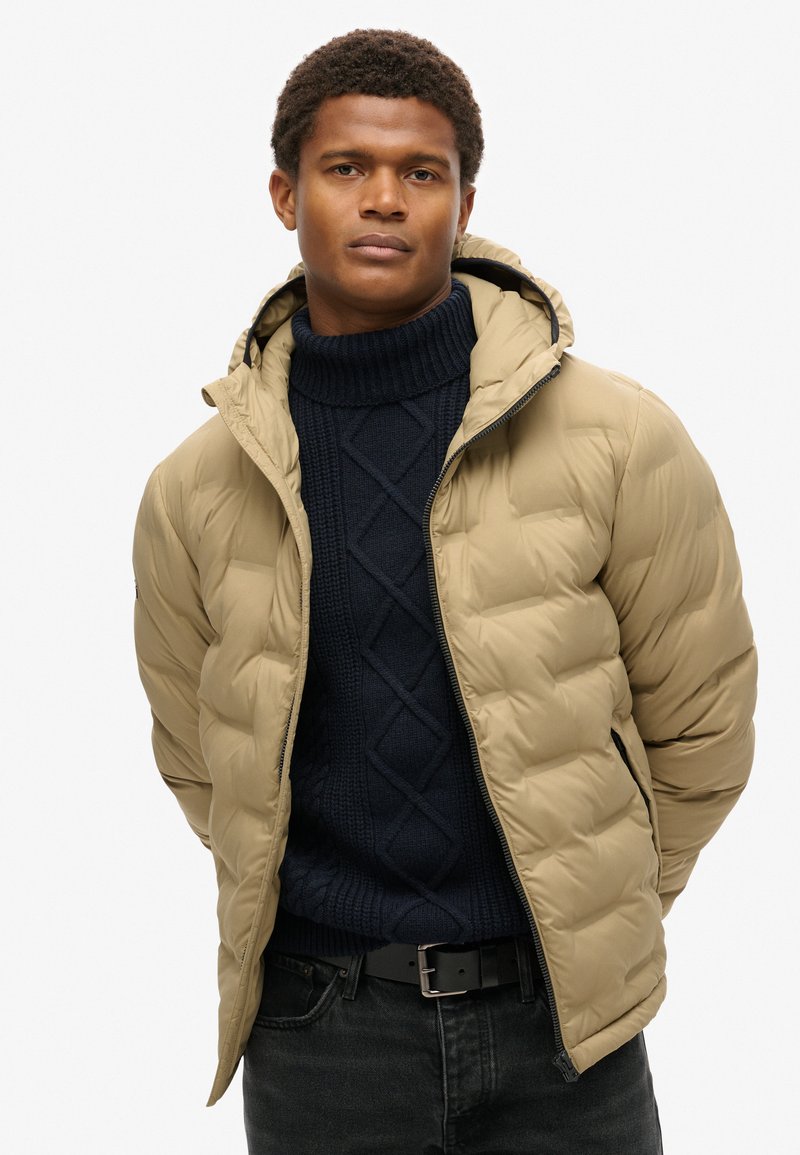 Chaqueta de plumas beige con capucha y textura acolchada, llevada sobre un suéter de punto azul marino. Se pueden ver un cinturón negro y jeans oscuros.