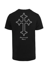 Zwarte katoenen t-shirt met een wit omrand kruis op de achterkant en de tekst "Memento Mori" onder het ontwerp. Korte mouwen, normale pasvorm.