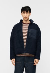 Νεαρός άνδρας με ναυτικό fleece μπουφάν με φερμουάρ σε τσέπη στο στήθος και κεντημένο λογότυπο, ριγέ πουκάμισο και σκούρο παντελόνι από τζιν.