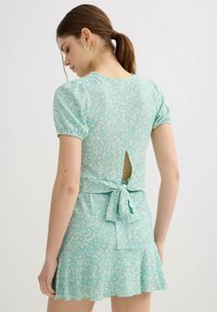 Mujer joven con coleta que lleva un vestido azul claro con estampado floral, mangas abullonadas, abertura en la espalda en forma de gota y lazo atado en la cintura.