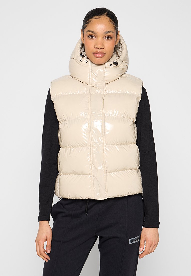 Gepolsterte Weste in glänzendem Beige, mit hohem Kragen und Kapuze. Reißverschluss mit Kordelzügen, über einem schwarzen Langarmoberteil getragen.