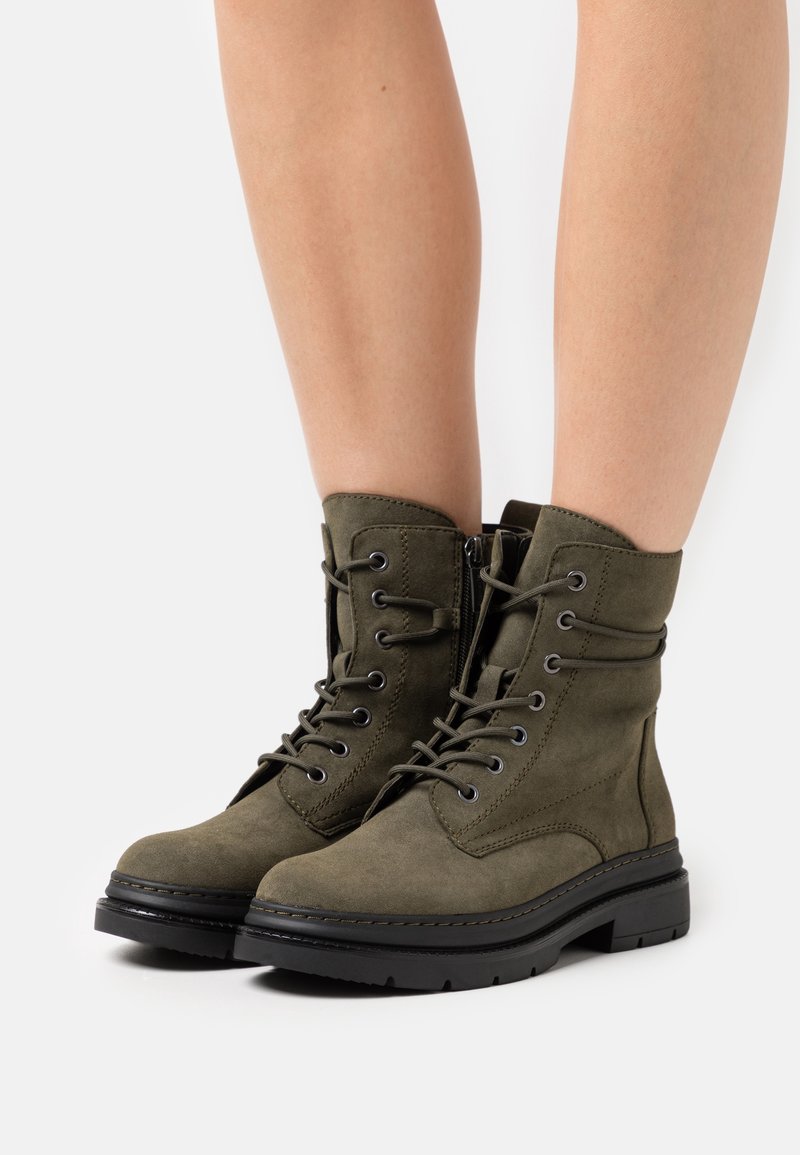 Paire de bottines en daim vert olive à lacets avec fermeture éclair latérale, portées sur des jambes nues contre un fond blanc.