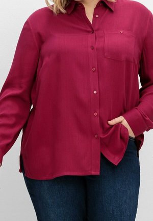 Chemise bordeaux à boutons, manches longues, tissu texturé, poche poitrine unique et fentes sur les côtés, associée à un jean denim foncé.