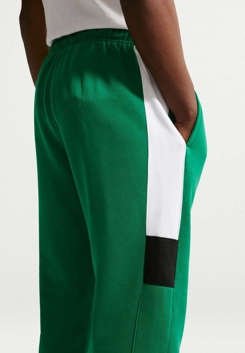 Persona che indossa pantaloni della tuta verdi con strisce verticali a blocchi di colore bianco e nero e mano in tasca, mostrata dalla vita in giù.