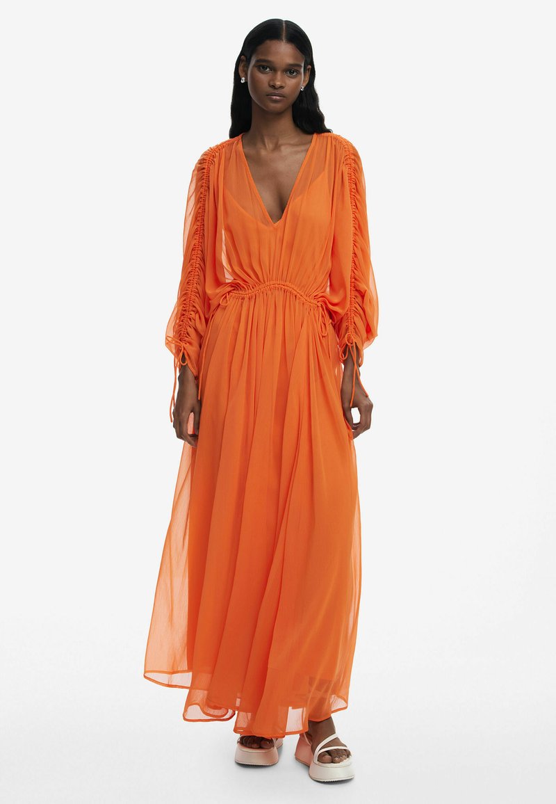 Maxiklänning i livlig orange chiffon, med djup V-ringning, samlad midja, puffärmar och flödande kjol. Bärs med platåsandaler.