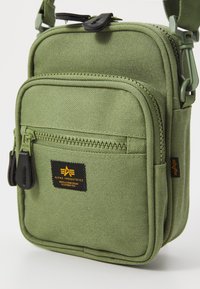 Borsa a spalla in tessuto verde oliva con due scomparti con zip, caratterizzata da un'etichetta logata nera e una superficie texturizzata per la durabilità.