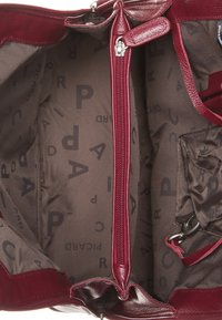 Burgunderfarbene Lederhandtasche mit einem Reißverschlussfach, einem Innenfutter aus Stoff mit wiederholtem Branding und einer glatten Außenfläche.