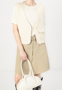 Chaleco beige acolchado de corte relajado, con dos bolsillos frontales, llevado sobre una blusa blanca texturizada y una falda beige, sosteniendo un bolso blanco.