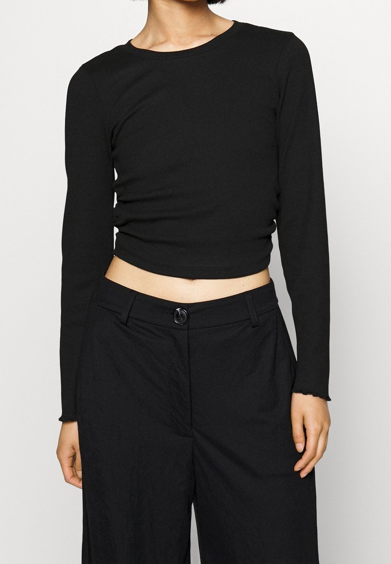 Černý dlouhý rukáv zkrácený top z měkké, žebrované látky s kulatým výstřihem a nedokončeným lemem, kombinovaný s černými kalhotami s vysokým pasem.