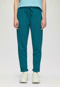 Tealowe joggery z elastycznym pasem i sznurkiem, z dwoma kieszeniami po bokach i zwężanym krojem nogawek. Połączone z białymi trampkami.