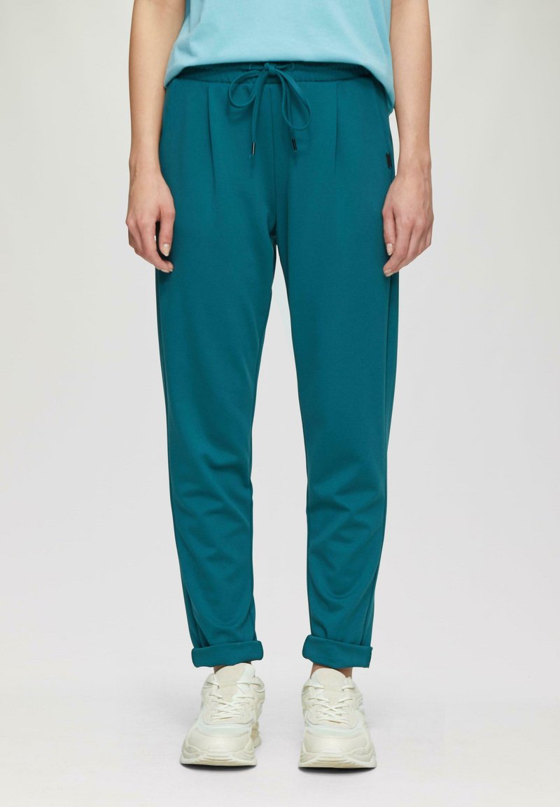 Tealowe joggery z elastycznym pasem i sznurkiem, z dwoma kieszeniami po bokach i zwężanym krojem nogawek. Połączone z białymi trampkami.