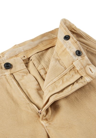 Pantalones beige con una cremallera abierta, dos botones negros y costuras visibles en la cinturilla sobre un fondo blanco.