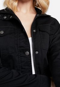 Blondýna v černé bundě přes bílý top, bunda částečně rozepnutá s kovovými patentkami a viditelnými náprsními kapsami.