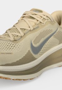 Ανοιχτό μπεζ αθλητικό παπούτσι Nike με γκρι λογότυπο Swoosh, επάνω μέρος από δίχτυ, επένδυση για τον αστράγαλο και μαλακή σόλα ορατή από το πλάι.