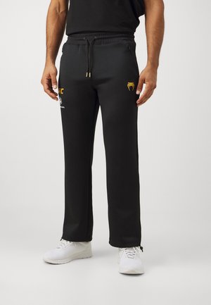 Mann trägt schwarze UFC-Jogginghose mit goldenem Schlangenlogo am rechten Oberschenkel und weiße Turnschuhe, steht vor neutralem Hintergrund.