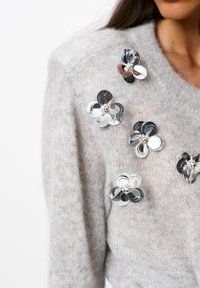 Lichtgrijze gebreide trui versierd met metallic bloemapplicaties in zilver, zwart en brons, met parelaccenten in het midden.