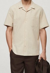 Korte mouwen beige shirt van zachte stof, met een ingesneden kraag, vijf knopen en een linkse borstzak. Ggedragen met donkerbruine broek.