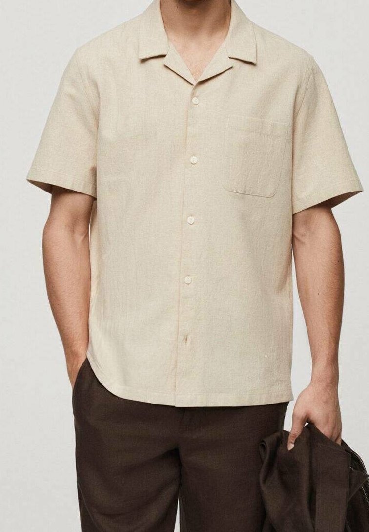 Korte mouwen beige shirt van zachte stof, met een ingesneden kraag, vijf knopen en een linkse borstzak. Ggedragen met donkerbruine broek.