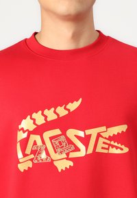 Felpa rossa con un grande logo in oro a forma di coccodrillo e testo. Il materiale è liscio con un collo a coste. I dettagli di design degni di nota includono un motivo testurizzato.