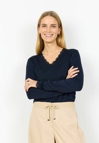 Tmavomodrý top s dlhým rukávom a výstrihom do V, s jemnou čiernou krajkou a hladkou textúrou. Kombinovaný s ľahkými béžovými nohavicami.