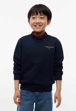 Ein lächelnder Junge mit kurzen schwarzen Haaren trägt ein marineblaues Tommy Hilfiger Sweatshirt und blaue Jeans und steht vor einem schlichten weißen Hintergrund.