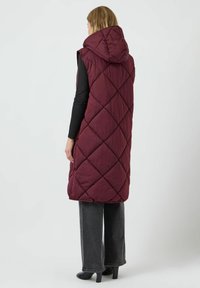 Burgundy, vadderad ärmlös väst med huva, med ett diamantmönster. Kombinerad med svart långärmad topp och grå vida byxor.