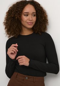 Black ribbed long-sleeve top with a rounded neckline. The model has a textured, curly hairstyle and is wearing a brown skirt with a button.